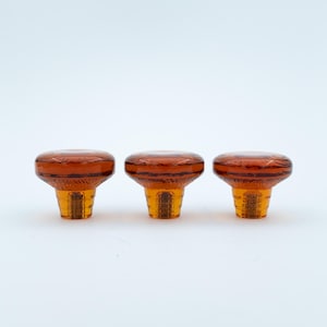 May include: Three amber-coloured glass knobs with a circular, flat top and a textured, tapered base. Each knob features a translucent, warm orange hue with a smooth, polished surface on the top and a ribbed, faceted design on the lower section.