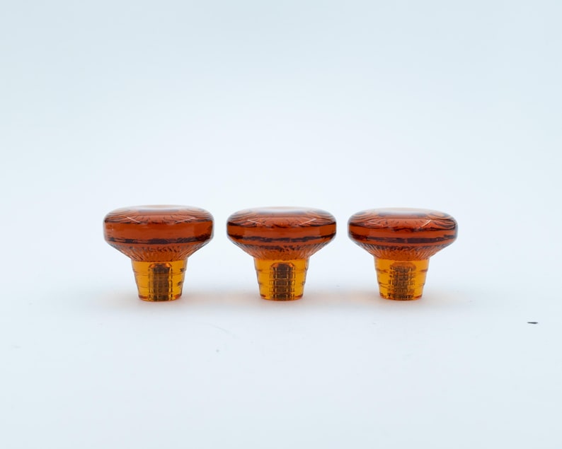 May include: Three amber-coloured glass knobs with a circular, flat top and a textured, tapered base. Each knob features a translucent, warm orange hue with a smooth, polished surface on the top and a ribbed, faceted design on the lower section.