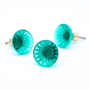 Könnte beinhalten: Drei türkisfarbene Glasschrankknöpfe mit facettiertem Design. Jeder Knopf hat einen messingfarbenen Sockel und eine Schraube zur Befestigung. Die Knöpfe sind auf weißem Hintergrund angeordnet und zeigen ihre Farbe und ihr Design.