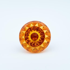 May include: A round, amber-colored glass knob with a starburst design. The knob features a central, raised circle surrounded by concentric rings of triangular and circular patterns. The glass has a glossy finish.