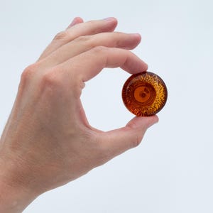 May include: A small, circular, amber-coloured glass object with a textured, sunburst-like pattern radiating from the centre. The piece is translucent, allowing light to pass through, and features a smooth, rounded edge, presenting a clear, polished finish.