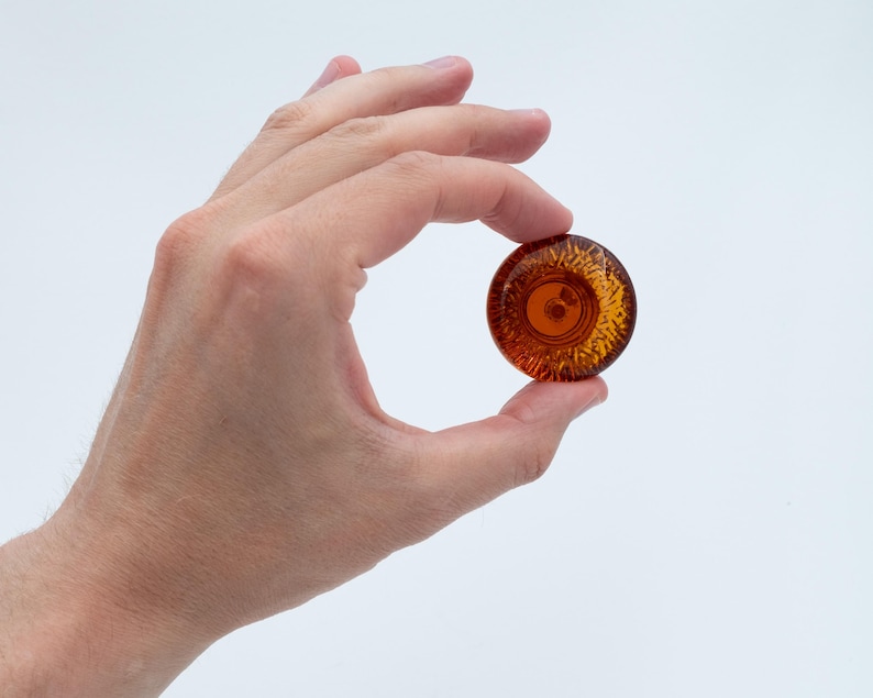 May include: A small, circular, amber-coloured glass object with a textured, sunburst-like pattern radiating from the centre. The piece is translucent, allowing light to pass through, and features a smooth, rounded edge, presenting a clear, polished finish.