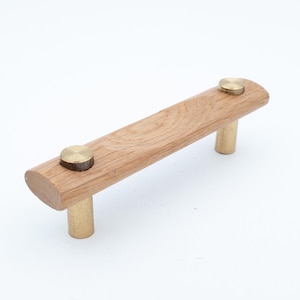 Peut inclure: Une poignée de meuble composée d'une barre horizontale en bois brun clair avec un grain visible. La barre est fixée à deux montants cylindriques en métal couleur laiton, chacun surmonté d'un capuchon plat et circulaire en métal laitonné.