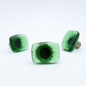 Op de afbeelding: Drie decoratieve flessenstoppers met een vierkante, transparante groene glazen bovenkant en een donkergroen centrum. De stoppers hebben een houten basis. Het ontwerp lijkt op een oog. De stoppers staan op een witte achtergrond.