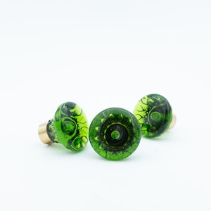 May include: Three round cabinet knobs made of translucent green glass with a dark, swirling internal pattern. Each knob features a smooth, domed surface and is attached to a cylindrical brass-colored metal base for mounting purposes.