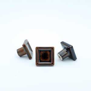 May include: Three brown square cabinet knobs with a dark centre and a light-coloured stem. The knobs are made of ceramic and have a glossy finish. The knobs are arranged on a white background.