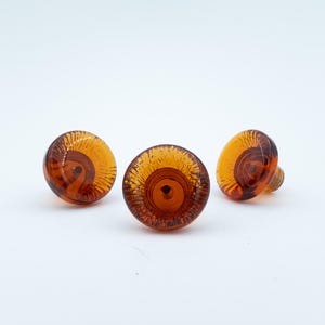 May include: Three round cabinet knobs made of translucent amber-coloured glass. Each knob features a circular, swirling pattern radiating from a central point, with a smooth, domed surface and a threaded metal attachment point visible on the back.