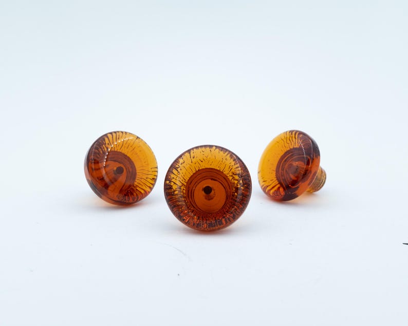 May include: Three round cabinet knobs made of translucent amber-coloured glass. Each knob features a circular, swirling pattern radiating from a central point, with a smooth, domed surface and a threaded metal attachment point visible on the back.