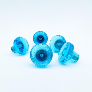 May include: Five clear blue glass cabinet knobs. Each knob has a round, textured face with a central indentation and a cylindrical stem. The knobs are arranged on a white background.