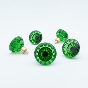 Può includere: Cinque pomelli per armadietti in vetro verde smeraldo con superfici sfaccettate e steli in ottone sono disposti su uno sfondo bianco. I pomelli hanno un design circolare con un cerchio scuro centrale.