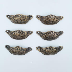 May include: Six semi-circular metal drawer pulls in an antique bronze finish. Each pull features a raised, symmetrical floral and leaf pattern with a central fan-like motif. The hardware includes two small circular screw holes on each side.