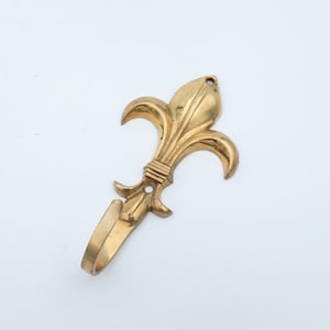 May include: A gold-colored metal wall hook shaped like a fleur-de-lis. The hook features a curved bottom section for hanging items and two small circular holes at the top and center for mounting purposes, with a smooth, polished metallic finish.