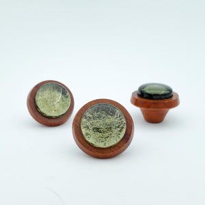May include: Three wooden cabinet knobs with a circular design. The knobs have a dark wood base and a clear, textured glass-like center with a green hue. One knob has a flat top, while the other two have a rounded top.