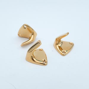 May include: Three gold-colored, triangular hooks with a curved design. Each hook has two small holes for mounting. The hooks are made of a shiny, metallic material and are set against a white background.