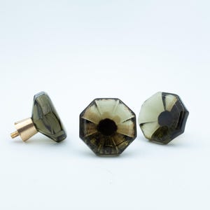 May include: Three octagonal cabinet knobs made of translucent dark grey glass with faceted edges. Each knob features a central dark circular detail and is attached to a cylindrical brass-coloured metal base with a threaded screw for installation.