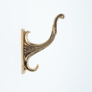 May include: A decorative brass wall hook with an ornate, curved design. The hook features a rectangular base and a curved arm with a small, decorative curl at the end. The hook is a warm, golden color and is mounted on a white background.