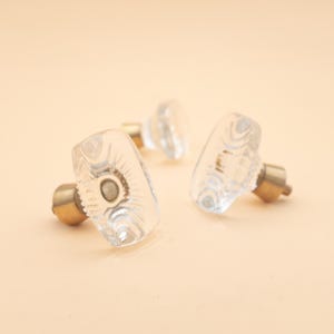 May include: Three clear glass cabinet knobs with brass hardware. The knobs have a rectangular shape with rounded edges and a decorative swirl pattern. The brass hardware is cylindrical. The knobs are on a light peach background.