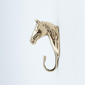 Könnte beinhalten: Ein metallischer, goldfarbener Wandhaken in Form eines Pferdekopfes. Das Design zeigt ein detailliertes Pferde-Profil mit einer fließenden Mähne, die sich nach unten erstreckt und an der Basis einen geschwungenen Haken bildet, der als Aufhängung dient.