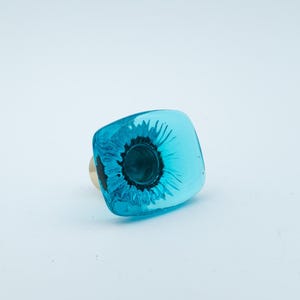 Könnte beinhalten: Ein türknopf aus türkisblauem Glas mit einem Blumendesign. Der Knopf hat eine quadratische Form und eine glänzende Oberfläche.