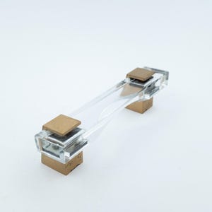 May include: A cabinet handle featuring a clear, transparent acrylic bar connected to two square, brushed gold-toned metal mounting bases. The design includes clear acrylic end caps that secure the central bar to the metallic square supports.