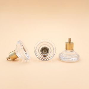 May include: Three clear glass cabinet knobs with a faceted design and brass fittings. The knobs are displayed on a light beige surface, highlighting their decorative details, suitable for home decor projects.