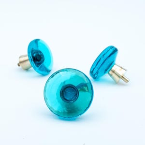 Peut inclure: Trois boutons de meuble en verre turquoise avec des fixations en laiton. Les boutons sont en forme de disque et disposés sur un fond blanc. Le verre est réfléchissant et les fixations en laiton ajoutent une touche d'élégance.