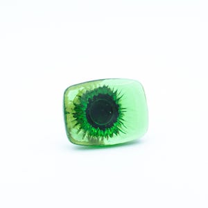 May include: A rectangular, green glass object with a detailed, dark green and black eye design. The eye is centered within the clear glass, creating a unique decorative piece. The object is set against a white background.