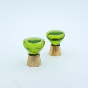 May include: Two green glass knobs with a rounded top and a brass base. The knobs are a vibrant green colour and have a slightly textured appearance. The brass base adds a touch of elegance. These knobs are likely for furniture or cabinetry.