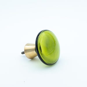 Pode incluir: Puxador decorativo para armário com um oval de vidro verde brilhante e uma base de latão. O puxador tem uma borda preta ao redor do vidro verde. A ferragem é de cor latão e é fixada a um parafuso.