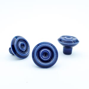 May include: Three navy blue ceramic cabinet knobs with a glossy finish. Each knob features a circular design with concentric rings and a small, rounded stem. The knobs are set against a white background.