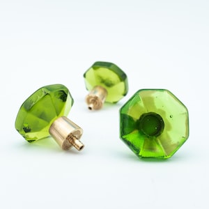 Pode incluir: Três botões de armário de vidro verde com bases de metal dourado. Os botões são facetados e têm uma forma geométrica.
