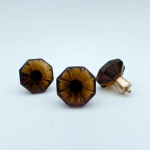 May include: Three octagonal amber glass cabinet knobs with brass hardware. The knobs have a faceted design and a dark center, creating a vintage aesthetic. Perfect for adding a touch of elegance to furniture.