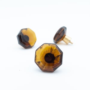 Könnte beinhalten: Drei bernsteinfarbene, facettierte Glasschrankknöpfe mit Messingbeschlägen. Die Knöpfe haben eine geometrische, achteckige Form und eine dunkle Mitte. Die Knöpfe sind vor einem weißen Hintergrund angeordnet.