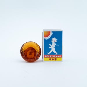 May include: A small, round, amber-coloured glass object with a textured, bubbly surface next to a rectangular matchbox. The matchbox features a blue background, a white silhouette of a child, a yellow sun, and the text SOLSTICKAN with three stars.