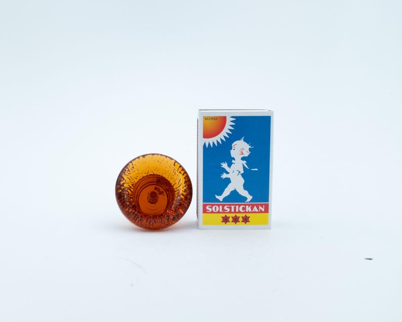 May include: A small, round, amber-coloured glass object with a textured, bubbly surface next to a rectangular matchbox. The matchbox features a blue background, a white silhouette of a child, a yellow sun, and the text SOLSTICKAN with three stars.