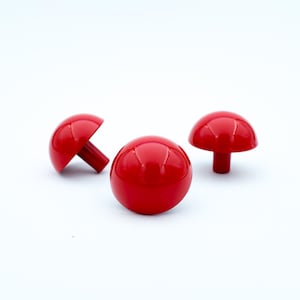 May include: Three red mushroom-shaped cabinet knobs. Each knob has a rounded top with vertical grooves and a cylindrical stem. The knobs are arranged on a white surface.