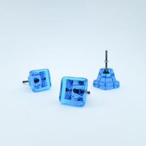 May include: Three blue glass cabinet knobs with square facets. Two knobs are lying down, and one is standing up. The knobs have a dark metal screw. The knobs are a bright, transparent blue color.