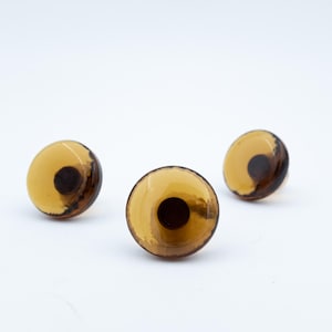 May include: Three amber-colored cabinet knobs with a dark brown center and a glossy finish. The knobs are round and have a unique, swirled design. They are set against a plain white background.