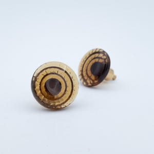 Peut inclure: Une paire de boucles d'oreilles à clous rondes présentant un dôme en verre translucide de couleur ambre avec un centre circulaire brun foncé et un motif métallique doré décoratif rayonnant vers l'extérieur en cercles concentriques.