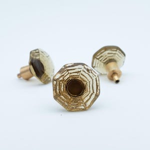 Puede incluir: Tres tiradores de armario de cristal vintage. Los tiradores son de color ámbar claro con una base de latón. Cada tirador tiene forma octogonal y un patrón similar a una telaraña.