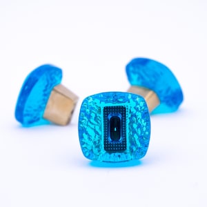 Puede incluir: Tres pomos de armario de vidrio azul con forma cuadrada y base de latón. Los pomos tienen una superficie texturizada y un pequeño cuadrado negro en el centro.