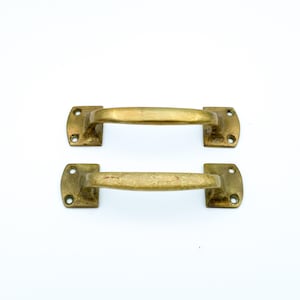 May include: Two antique brass cabinet pulls. Each pull has a rectangular base with rounded corners and a curved handle. The brass has a warm, aged patina, suggesting vintage or reclaimed hardware. The pulls are ideal for furniture restoration.