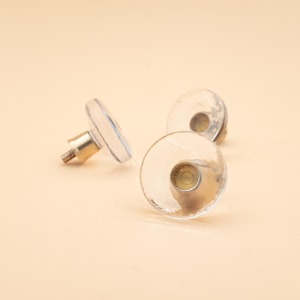 May include: Three clear glass cabinet knobs with brass stems on a light peach background. The knobs are circular with a textured surface. The brass stems are cylindrical with a threaded end. The knobs are suitable for furniture.