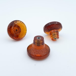 May include: Three amber-coloured glass knobs with a circular, flat top and a tapered base. Each knob features a textured, radial pattern radiating from the centre of the top surface and a metal threaded insert at the base for mounting purposes.
