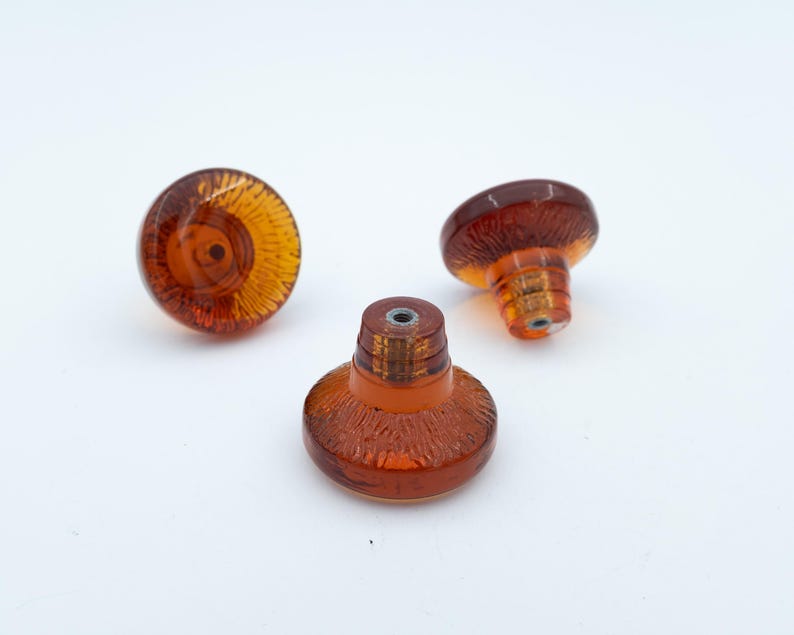 May include: Three amber-coloured glass knobs with a circular, flat top and a tapered base. Each knob features a textured, radial pattern radiating from the centre of the top surface and a metal threaded insert at the base for mounting purposes.