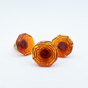 May include: Three amber-colored glass knobs with an octagonal shape and a faceted surface. Each knob features a central circular indentation and a metallic base, showcasing a translucent, warm orange hue that catches the light.