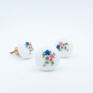 May include: Three round white ceramic cabinet knobs featuring a delicate floral pattern with blue, pink, and white flowers and green leaves. Each knob has a smooth, glossy finish and a metallic mounting screw attached to the back for installation.
