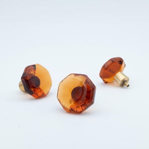 Puede incluir: Tres tiradores de armario de cristal facetado color ámbar con herrajes de latón. Los tiradores tienen forma octogonal y un tono cálido y transparente, lo que sugiere un estilo vintage. Los herrajes de latón añaden un toque de elegancia.