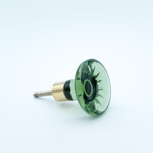 Peut inclure: Un bouton de meuble en verre vert avec une base en laiton et une vis argentée. Le bouton a une forme arrondie et légèrement bombée avec un motif décoratif à l'intérieur. La quincaillerie est conçue pour les meubles.