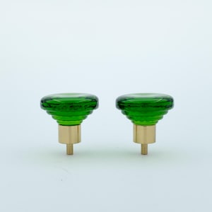 Può includere: Due pomelli per armadietti in vetro verde con un design a coste. I pomelli hanno una base in ottone e una vite per l'installazione. I pomelli sono di un vivace colore verde e sono su uno sfondo bianco.